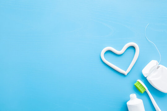 Toothbrush, white tube of toothpaste, container of dental floss on blue table background. Pastel color. Heart shape created from paste. Love healthy teeth concept. Empty place for text. Top down view.