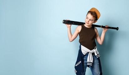 Sly little schoolboy stands near the wall looking aside holding baseball bat on shoulders. Three...
