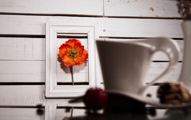 The poppy flower in a frame for a picture and the coffee cup with cinnamon on the brown wodden table and white vase with flowers for coffee time.