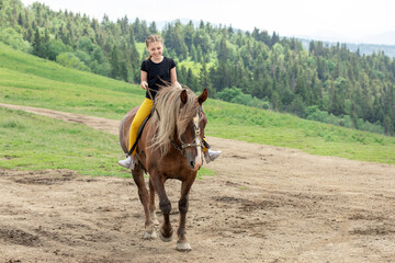 Happy teenager girl riding a horse on a mountainous area. The girl spends her leisure time with a smile. She likes this hobby.