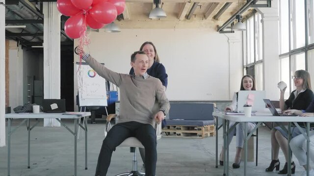 Colleagues congratulate the young manager. They ride him in an office chair, give him red balloons, explode crackers with confetti. Business team dancing and celebrating. Corporate. Office life