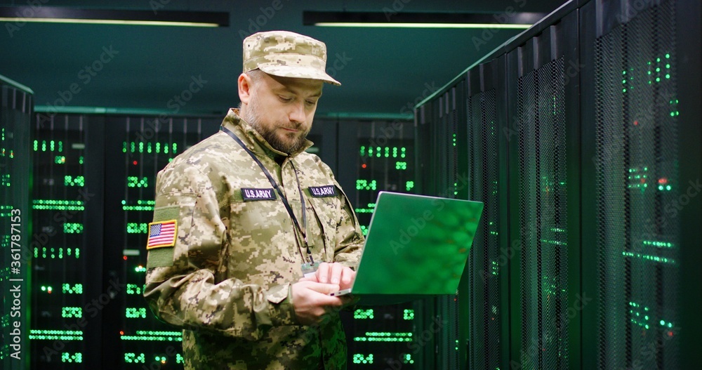 Wall mural us army male caucasian officer in uniform and cap stading at server wih secret data and using laptop