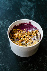 Take Away Vegan Food Acai Bowl with Almond Milk, Peanut Butter, Banana, Buckwheat Granola, Chia Seeds and Chocolate Pieces in Carton Bowl.