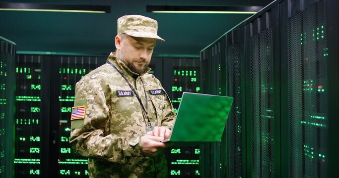 US army male Caucasian officer in uniform and cap stading at server wih secret data and using laptop computer to check the information. Military monitoring service room. Army networking concept.