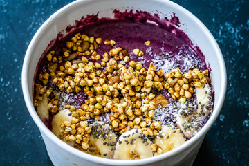 Take Away Vegan Food Acai Bowl with Almond Milk, Peanut Butter, Banana, Buckwheat Granola, Chia Seeds and Chocolate Pieces in Carton Bowl.
