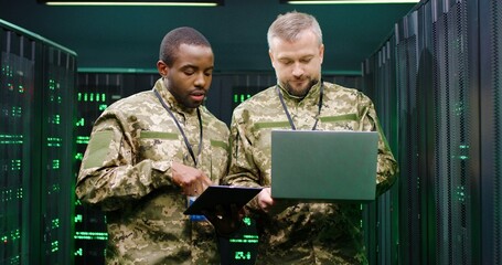 Fototapeta premium Mixed-races army men in camouflage talking and discussing some secret information in server room. African American and Caucasian male co-workers of military forces with tablet and laptop computer.