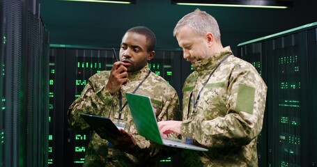 Fototapeta premium Multiethnic male army officers with tablet device and laptop computer checking secret data in server room and passing via walkie-talkie. African American and Caucasian men talking at digital facility.