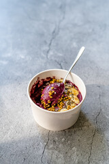 Take Away Vegan Food Acai Bowl with Almond Milk, Peanut Butter, Banana, Buckwheat Granola, Chia Seeds and Chocolate Pieces in Carton Bowl.