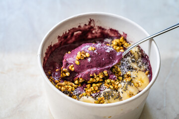 Take Away Vegan Food Acai Bowl with Almond Milk, Peanut Butter, Banana, Buckwheat Granola, Chia Seeds and Chocolate Pieces in Carton Bowl.