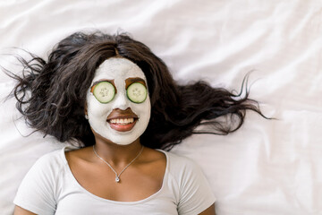Top-view of smiling African woman making spa procedures with mud facial mask and fresh cucumber slices on eyes, laying on bed. Wellness and skincare concept