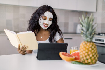 Portrait of young African woman with mud facial mask on her face, sitting at the table with fresh fruits in modern kitchen and working or studying using ipad tablet.