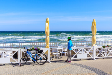 Young woman cyclist with bike standing on promenade and looking at white sand beach with blue sea in Kolobrzeg town, Baltic Sea coast, Poland