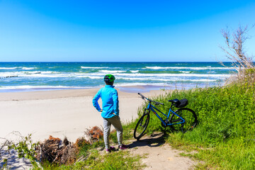 Young woman cyclist standing on green sand dune and looking at beautiful white sand beach with blue sea near Kolobrzeg, Baltic Sea coast, Poland - obrazy, fototapety, plakaty