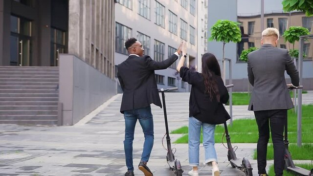 Back view of good-looking trendy happy young mates which holding in their hands electric scooters and giving high five each other on the urban street near beautiful building
