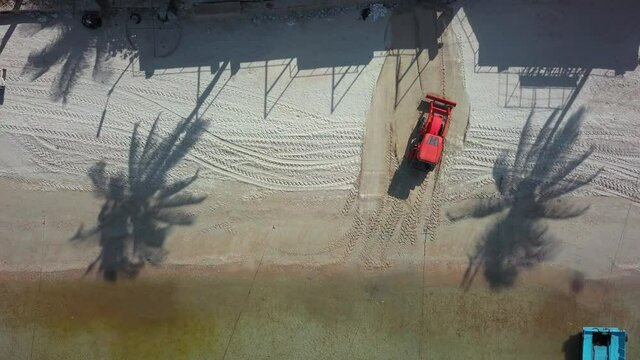 Overhead View Of Red Tractor Drive Away From Frame, Machine Cleaning A Sand Beach Near Coastline On A Sunny Day. Still Lock Down Static Video Shot