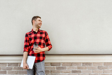 Young happy man freelancer is standing near a bricks wall and holding a coffee and a laptop in his hands