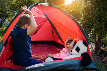 Two happy little kids, boy and girl brother and sister are lying and playing in a red camping tent  with their panda bear toy in the home yard on the grass.
