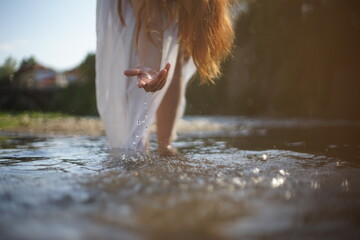 A girl/ young woman with red hair and a white dress. She is playing with water. The photo is taken in a rural scene. The weather is sunny, yet the light is soft and warm.