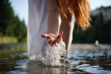 A girl/ young woman with red hair and a white dress. She is playing with water. The photo is taken in a rural scene. The weather is sunny, yet the light is soft and warm.