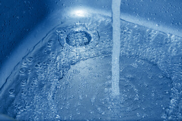 A stream of clean water running down the drain into the stainless steel sink in blue tones.