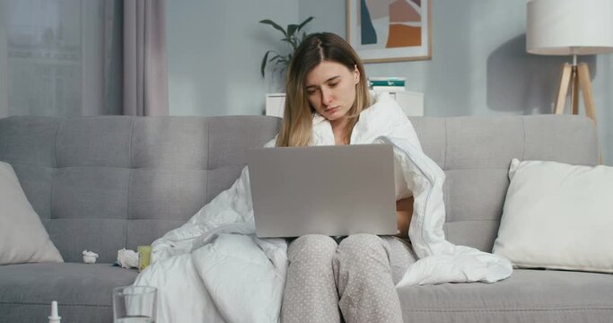 Caucasian young ill female using laptop while sitting on couch in blanket, caughing and feeling bad at home. Unhealthy girl tapping, typing and scrolling news on computer on sofa in living room.