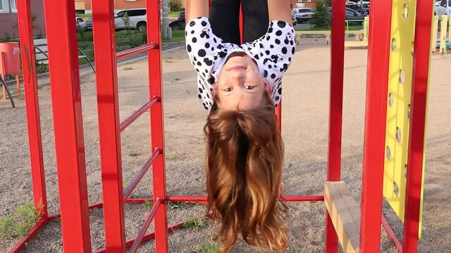 Happy girl child swinging upside down in the playground. Child plays on the horizontal bar and stairs.