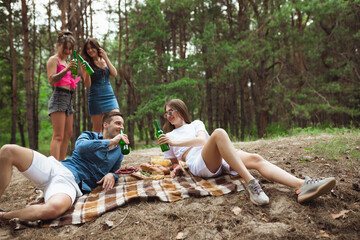 Happiness. Group of friends clinking beer bottles during picnic in summer forest. Lifestyle, friendship, having fun, weekend and resting concept. Looks cheerful, happy, celebrating, festive.