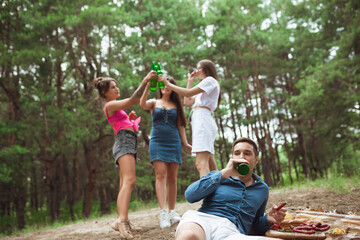 Obraz premium Togetherness. Group of friends clinking beer bottles during picnic in summer forest. Lifestyle, friendship, having fun, weekend and resting concept. Looks cheerful, happy, celebrating, festive.