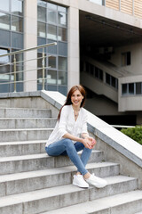 Attractive young woman in the city. Business lady standing near business center. Tired business woman with white shirt in blue jeans sitting relax on the street near city centre glass building