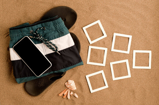 summer on the beach, black flip flops with trendy men's swimsuit and black screen smartphone, shells, frames mockup photo college on fine beach sand. Top view.