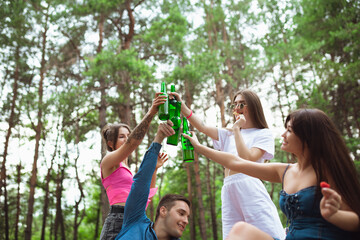 Fototapeta premium Youth. Group of friends clinking beer bottles during picnic in summer forest. Lifestyle, friendship, having fun, weekend and resting concept. Looks cheerful, happy, celebrating, festive.