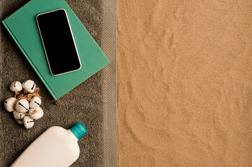 summer in the beach concept, cris towel on beach sand, on top of turquoise book with smartphone black screen, bottle of cream. beach sand .top view.
space for text