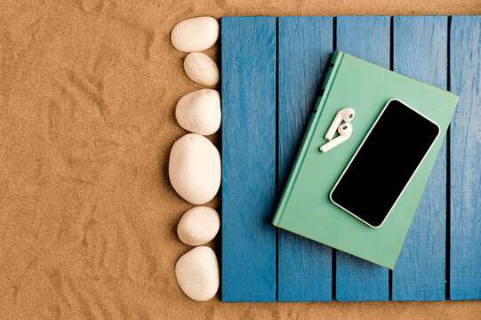 summer in the beach concept, blue wooden platform on beach sand, book with smartphone and headphones on it, white stones. beach sand background.top view