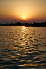 Deep calm dawn on the lake with rising sun over the hills. The Garda lake water mirror the warm colors of the sunshine, sky is cloudless and all is quite.