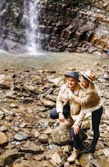 Young and beautiful couple at the mountain waterfall - Happy tourists visiting mountains. Lovestory. Tourists in hats. Military fashion.