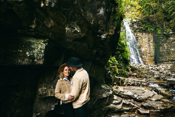 Young and beautiful couple at the mountain waterfall - Happy tourists visiting mountains. Lovestory. Tourists in hats. Military fashion.