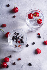 Fresh cold sparkling water drink with cherry, raspberry and currant berries in two transparent glass on stone concrete background, summer diet beverage, angle view