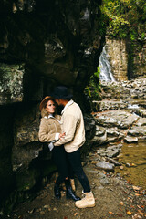 Young and beautiful couple at the mountain waterfall - Happy tourists visiting mountains. Lovestory. Tourists in hats. Military fashion.