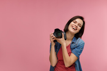 Portrait of young female photographer isolated on pink background with copy space. Photographer with professional camera taking photo in photo studio. Profession, occupation, self-employment.