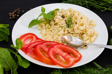 Bulgur, greens and vegetables in a plate and spoon on a black background. Close-up. Tasty and healthy vegetarian food.