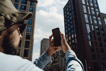Tourist man in hat travels around the city. A guy with a backpack and sunglasses. The traveler stopped to take a photo on phone. Hipster with a beard. Communication technology in an urban environment.