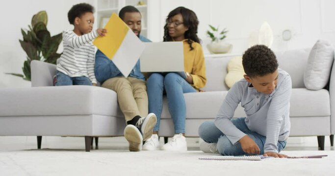 Small cute African American children playing and painting pictures with parents in cozy living room. Parents drawing with little son and daughter at home. Family spending time together.