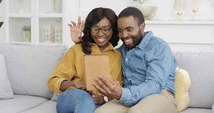 Young African American couple sitting on couch in living room and videochatting on tablet device. Male and female talking via webcam on computer at home on white sofa. Man and woman having videochat.