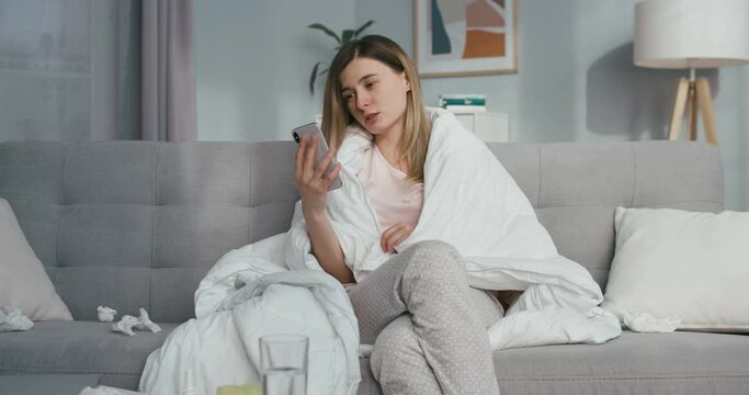 Ill young Caucasian woman with fever sitting on couch at home and videochatting on smartphone. Sick girl caughing and having videochat on mobile phone via webcam. Coronavirus isolation.