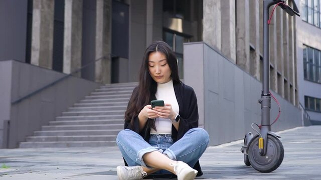 Agreeable satisfied young stylish asian girl sitting on the concrete ground near own e-scooter on the beautiful urban building background and using her smartphone