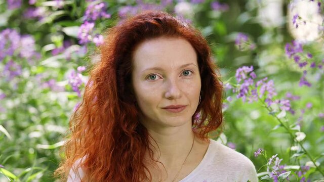 Portrait of a redhead young adult woman among flowers in a city park on a sunny summer day
