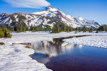 Volcanic mountain peak under a crisp blue sky rises behind a field of snow.  Calm water in the foreground reflects the natural scene.
