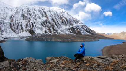Man sitting at the turquoise colored Tilicho lake in Himalayas, Manang region in Nepal. The world's highest altitude lake (4949m). Snow capped mountains around. Calm surface of the lake. Achievement