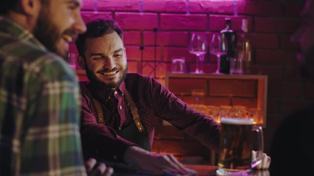 In a beer pub good looking bart enjoying the time at work smiling large he cleaning the bar table while other customers spending a good time at bar table