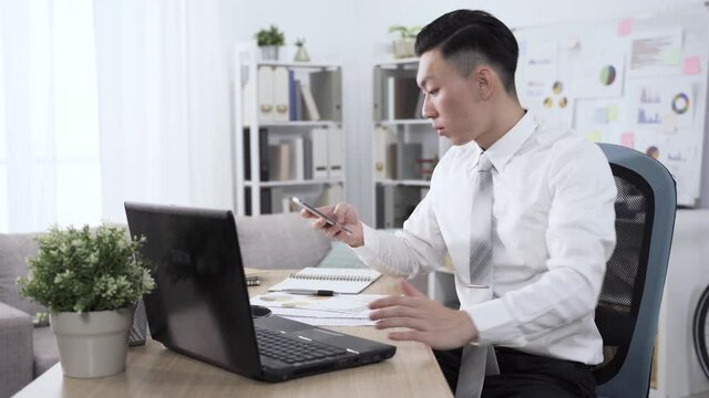 chinese guy sitting at desk typing is answering a phone. businessman leaning back comfortably in chair, putting hand in pocket and crossing legs while speaking on smartphone.